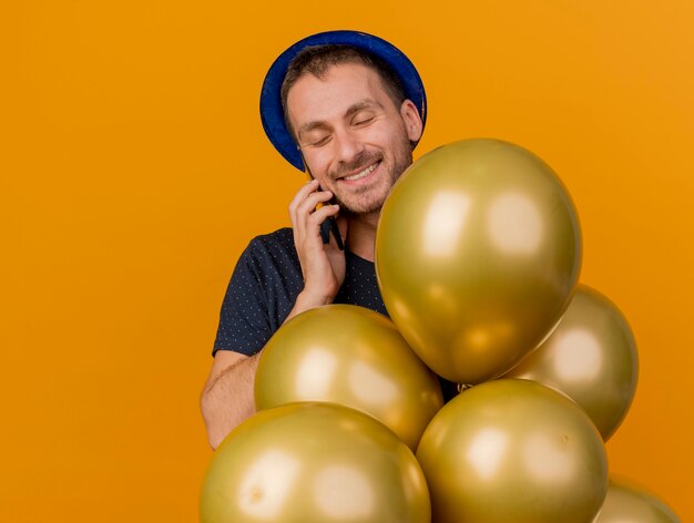 Sourire bel homme caucasien portant chapeau de fête bleu détient des ballons d'hélium parler au téléphone isolé sur fond orange avec espace de copie