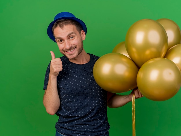 Photo gratuite sourire bel homme caucasien portant chapeau de fête bleu pouces vers le haut et détient des ballons d'hélium isolés sur fond vert avec espace de copie