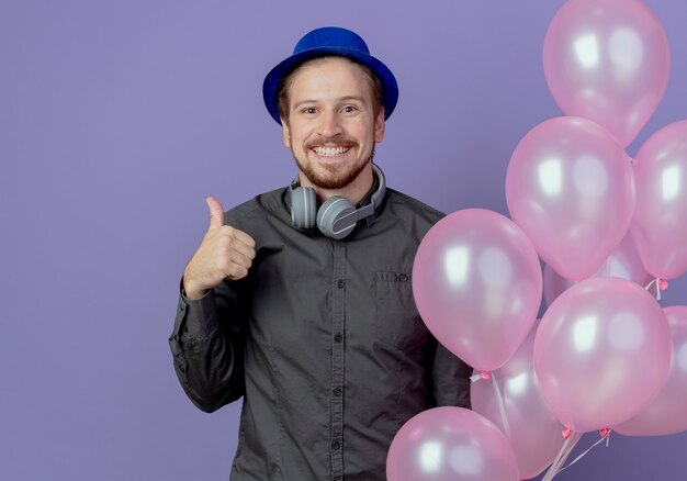 Sourire bel homme avec chapeau bleu et écouteurs sur le cou se dresse avec des ballons d'hélium pouces vers le haut isolé sur mur violet