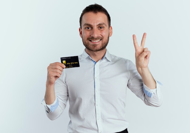 Sourire bel homme détient une carte de crédit et deux gestes avec la main isolé sur un mur blanc