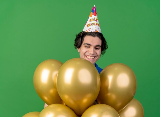Sourire bel homme portant une casquette d'anniversaire ressemble et se tient avec des ballons d'hélium isolés sur un mur vert