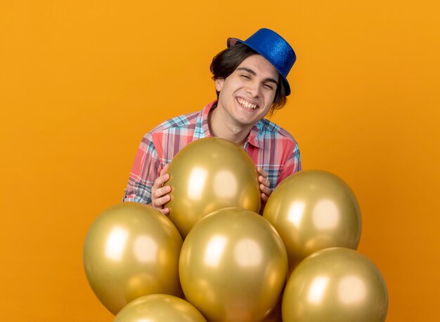 Sourire bel homme portant un chapeau de fête bleu se dresse avec des ballons d'hélium isolés sur un mur orange