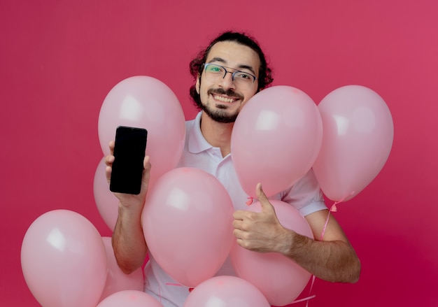 Sourire bel homme portant des lunettes debout derrière des ballons tenant le téléphone son pouce vers le haut isolé sur un mur rose