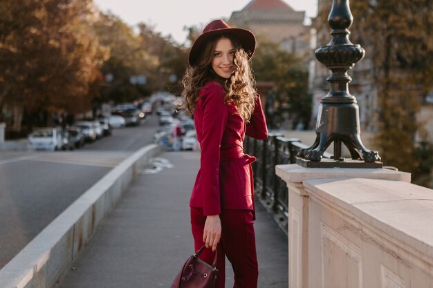 Sourire belle femme élégante en costume violet marchant dans la rue de la ville, tendance de la mode printemps été saison automne portant chapeau, tenant sac à main