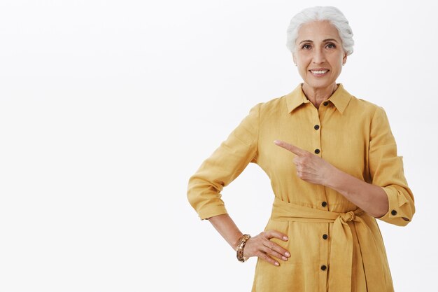 Sourire belle femme senior en manteau jaune doigt pointé vers la gauche, montrant la publicité