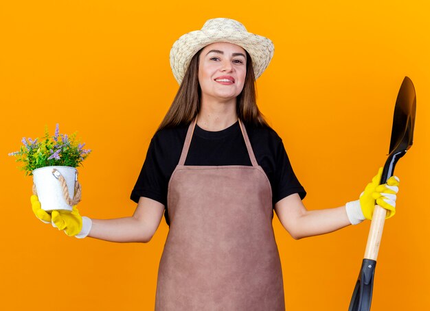 Sourire belle fille de jardinier portant uniforme et chapeau de jardinage avec des gants tenant la pelle avec fleur en pot de fleurs et répandre les mains isolées sur fond orange