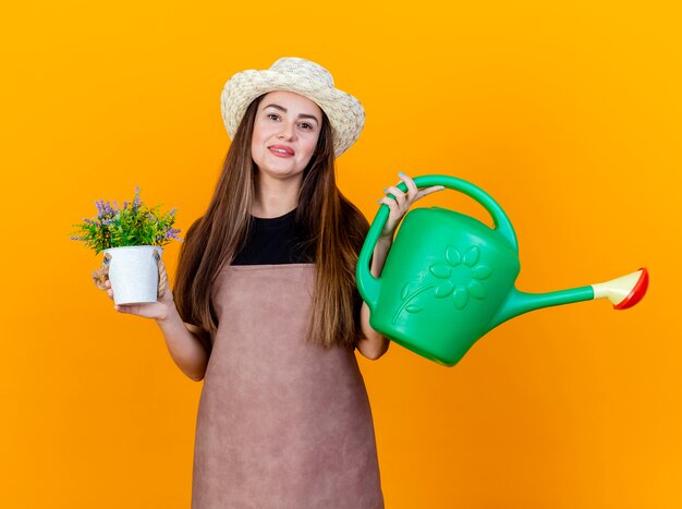 Sourire belle fille de jardinier en uniforme et chapeau de jardinage tenant un arrosoir avec fleur en flwerpot isolé sur fond orange