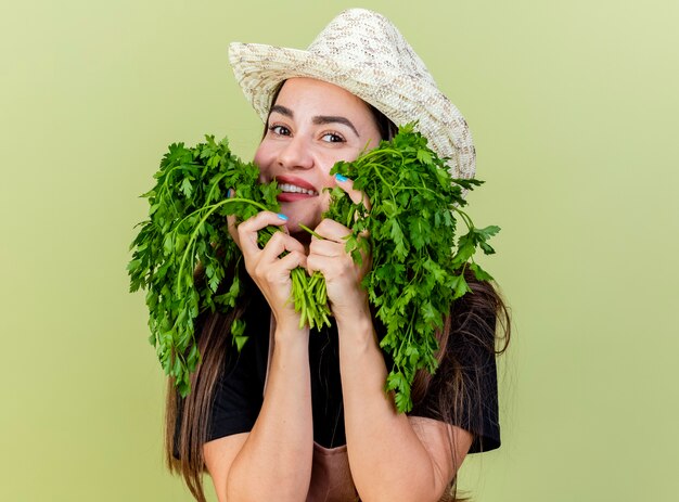 Sourire belle fille de jardinier en uniforme portant chapeau de jardinage mettant la coriandre sur les joues isolé sur fond vert olive