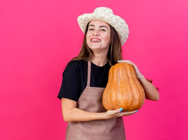 Sourire belle fille de jardinier en uniforme portant chapeau de jardinage tenant citrouille isolé sur fond rose