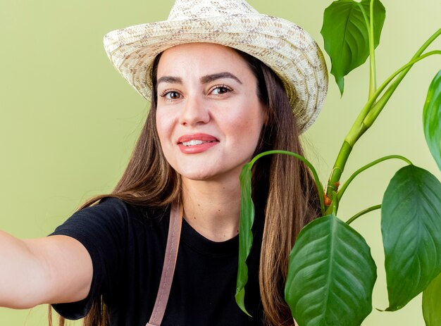 Sourire belle fille de jardinier en uniforme portant chapeau de jardinage tenant plante avec appareil photo isolé sur vert olive