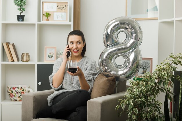 Sourire à côté de belle fille le jour de la femme heureuse tenant un verre de vin parle sur le vin assis sur un fauteuil dans le salon