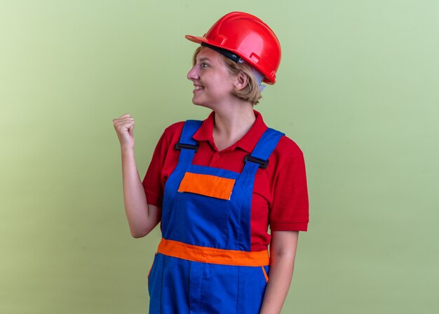 sourire à côté jeune constructeur femme en uniforme montrant oui geste isolé sur mur vert olive