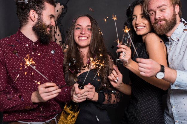 Sourire de dames et de gars tenant des feux de Bengale, une bouteille et un verre de boisson