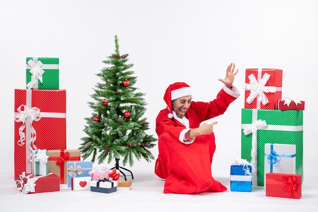 Sourire drôle de jeune homme habillé en père Noël avec des cadeaux et arbre de Noël décoré assis sur le sol en pointant quelque chose sur le côté gauche sur fond blanc