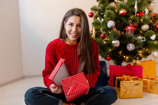 Sourire excité jolie femme en pull rouge assis à la maison à l'arbre de Noël déballage des cadeaux et coffrets cadeaux