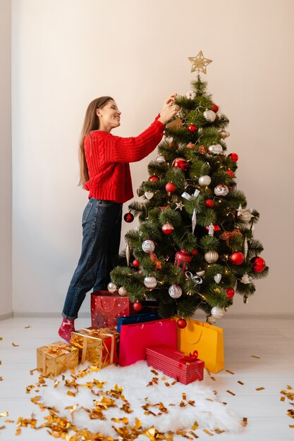 Sourire excité jolie femme en pull rouge debout à la maison décoration arbre de Noël entouré de cadeaux et coffrets cadeaux