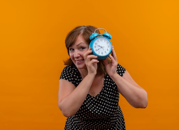 Sourire de femme d'âge moyen tenant un réveil et regardant derrière sur un mur orange isolé avec copie espace