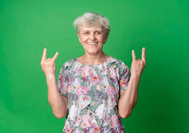 Sourire femme âgée gestes cornes avec deux mains isolés sur un mur vert