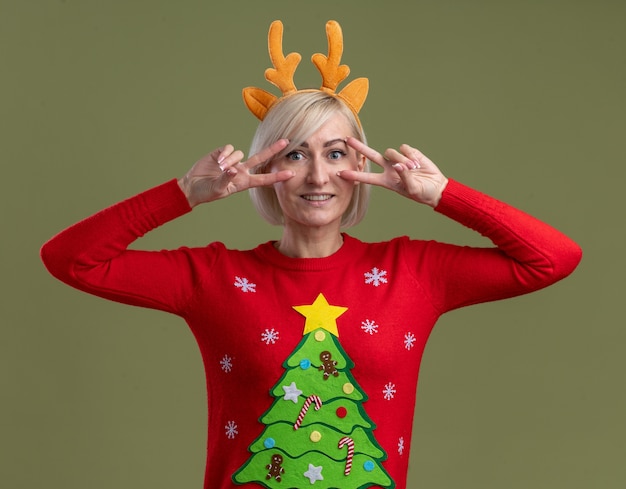 Sourire d'une femme blonde d'âge moyen portant un bandeau de bois de renne de Noël et un pull de Noël regardant la caméra montrant des symboles v-sign près des yeux isolés sur fond vert olive