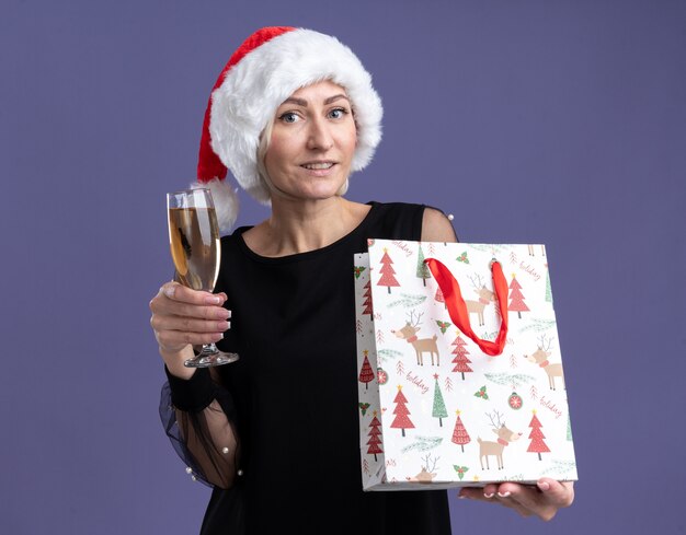 Sourire de femme blonde d'âge moyen portant un chapeau de Noël regardant la caméra tenant un verre de champagne et sac-cadeau de Noël isolé sur fond violet