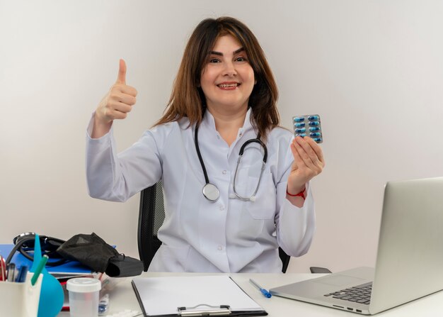 Sourire femme médecin d'âge moyen portant portant une robe médicale avec stéthoscope assis au bureau de travail sur un ordinateur portable avec des outils médicaux tenant une carte de crédit son pouce vers le haut sur un mur blanc