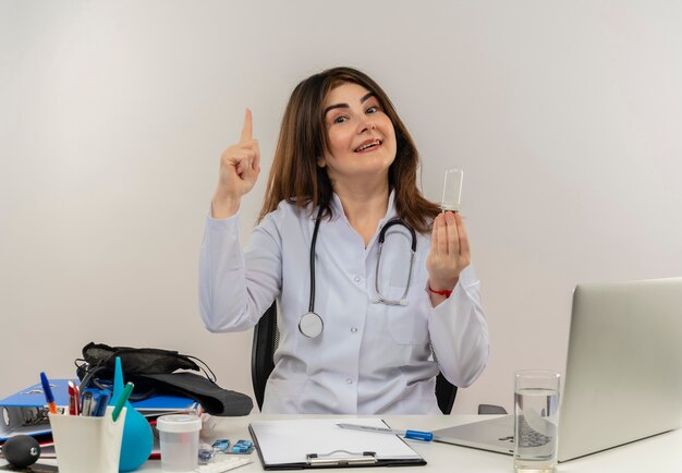 Sourire femme médecin d'âge moyen portant portant une robe médicale avec stéthoscope assis au bureau travailler sur un ordinateur portable avec des outils médicaux tenant une ampoule et pointer vers le haut sur un mur blanc