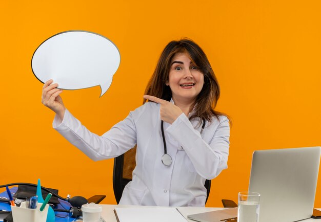 Sourire de femme médecin d'âge moyen portant une robe médicale avec stéthoscope assis au bureau de travail sur un ordinateur portable avec des outils médicaux tenant et des points pour discuter de la bulle sur le mur orange