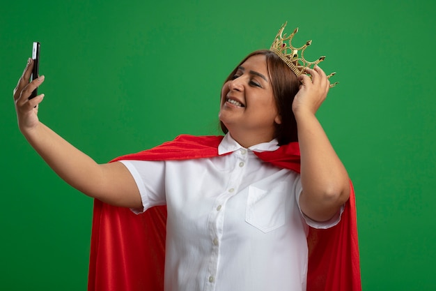 Sourire femme de super-héros d'âge moyen portant couronne prendre un selfie mettant la main sur la couronne isolé sur vert