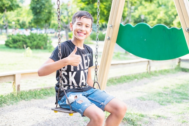 Sourire garçon assis dans la balançoire montrant le pouce en haut signe dans le parc