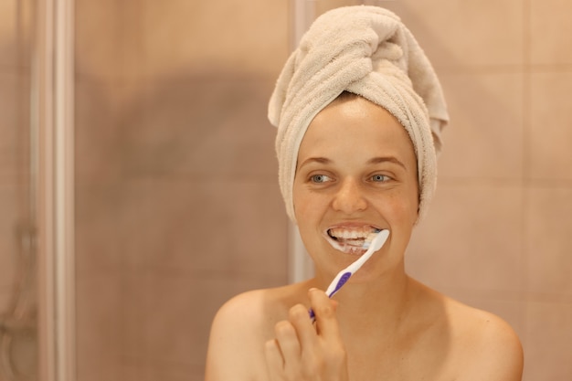 Sourire heureuse femme séduisante de bonne humeur se brosser les dents, avoir des procédures d'hygiène et de beauté à la maison dans la salle de bain, être enveloppée dans une serviette blanche.