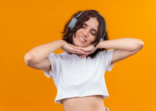 Sourire heureux jeune belle femme portant des écouteurs et mettant les mains sous son menton sur un mur orange isolé