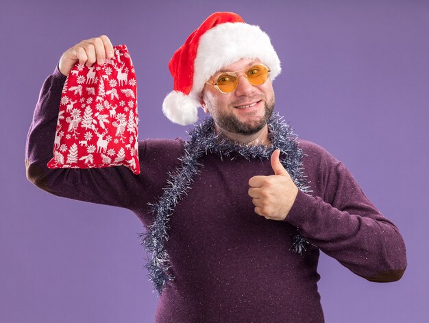 Sourire homme d'âge moyen portant bonnet de Noel et guirlande de guirlandes autour du cou avec des lunettes tenant le sac de cadeau de Noël montrant le pouce vers le haut isolé sur mur violet