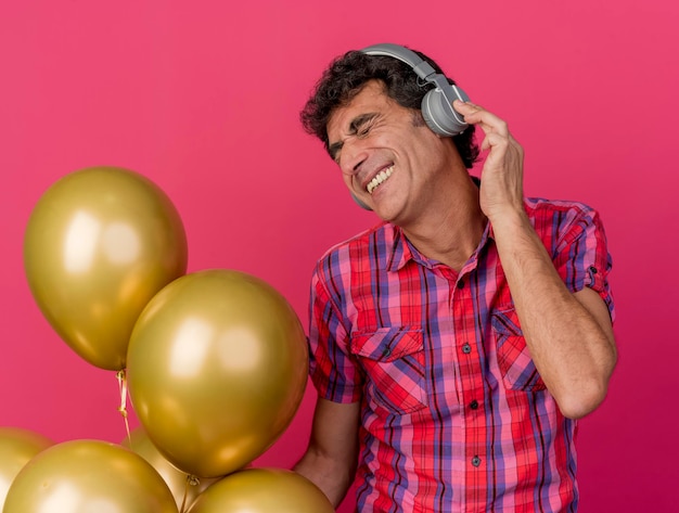 Sourire homme d'âge moyen portant des écouteurs tenant des ballons, écouter de la musique avec les yeux fermés isolé sur un mur rose