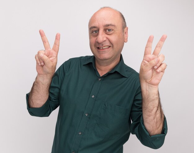 Sourire homme d'âge moyen portant un t-shirt vert montrant le geste de paix isolé sur un mur blanc