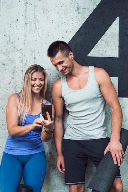 Sourire homme et femme regardant un téléphone portable contre le mur dans un club de fitness