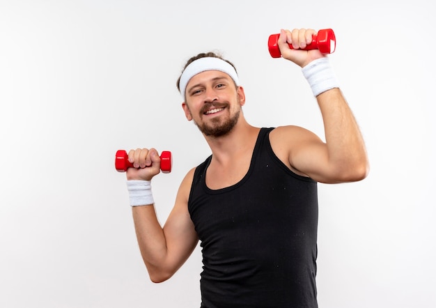 Sourire jeune bel homme sportif portant un bandeau et des bracelets tenant des haltères isolés sur un espace blanc