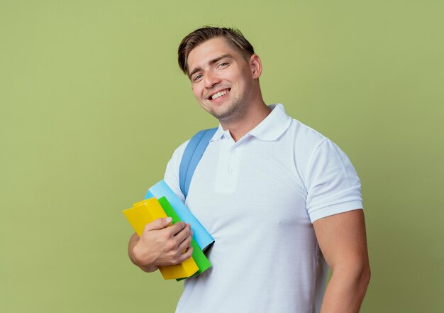 Sourire jeune étudiant beau mâle portant sac à dos tenant des livres isolés sur vert olive