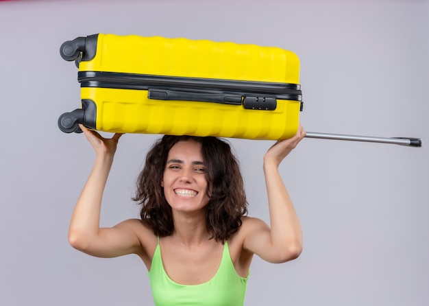 Photo gratuite sourire jeune femme belle voyageur mettant valise sur sa tête sur un mur blanc isolé