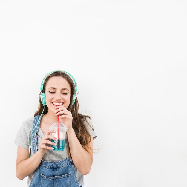 Sourire jeune femme écoutant de la musique sur le casque profiter de boire du jus sur fond blanc