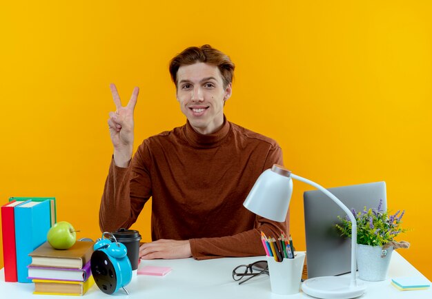 Sourire jeune garçon étudiant assis au bureau avec des outils scolaires geste de paix sur jaune