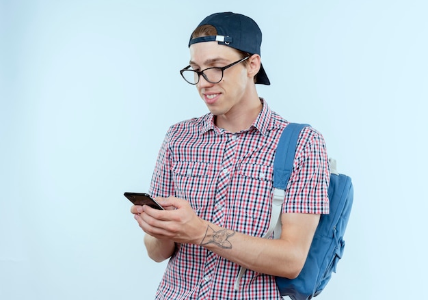 Sourire jeune garçon étudiant portant un sac à dos et des lunettes et une casquette tenant et regardant le téléphone sur blanc