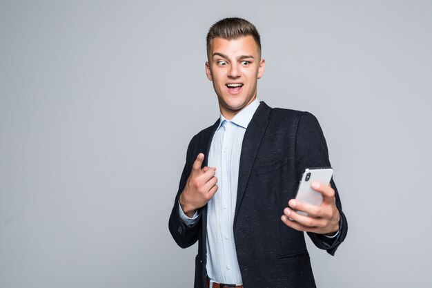 Sourire jeune homme a un appel vidéo sur un téléphone habillé en veste sombre en studio isolé sur mur gris