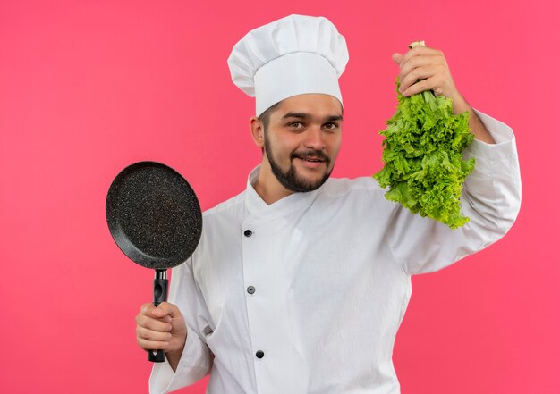 Sourire jeune homme cuisinier en uniforme de chef tenant poêle et laitue isolé sur espace rose