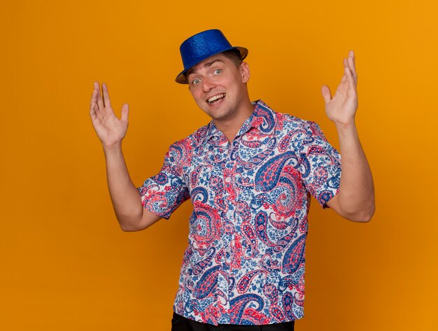 Sourire jeune mec de fête portant un chapeau bleu répandre les mains isolés sur orange