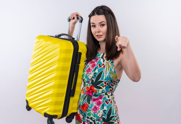 Sourire jeune voyageur femme vêtue d'une robe multicolore tenant un sac mobile et pointe vers l'avant sur un mur blanc
