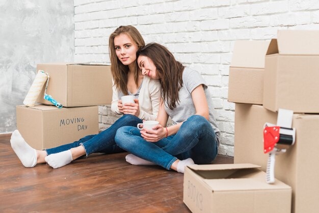 Sourire de jeunes femmes assis sur le sol, tenant des tasses à café à la main, assis entre les piles de boîtes en carton