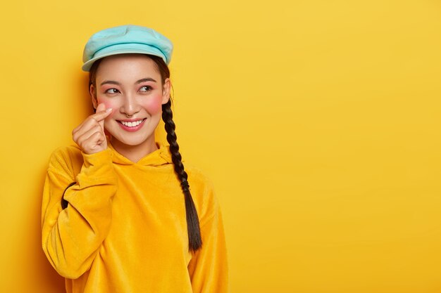 Sourire jolie femme Asain avec des nattes, a les joues rouges, fait un signe coréen, porte un béret et un sweat-shirt, a une expression de visage de rêve