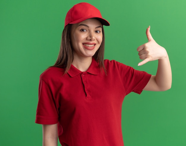 Sourire jolie femme de livraison en uniforme faisant des gestes accrocher signe lâche isolé sur mur vert avec espace copie