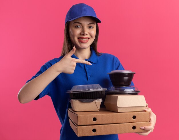 Sourire jolie femme de livraison en uniforme tenant et pointant sur le paquet de nourriture et les conteneurs sur les boîtes de pizza sur rose