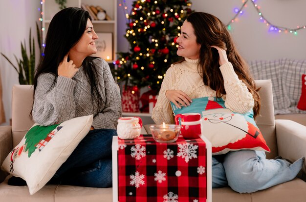 Sourire de jolies jeunes filles se regardent soulever leurs cheveux assis sur des fauteuils et profiter du temps de Noël à la maison